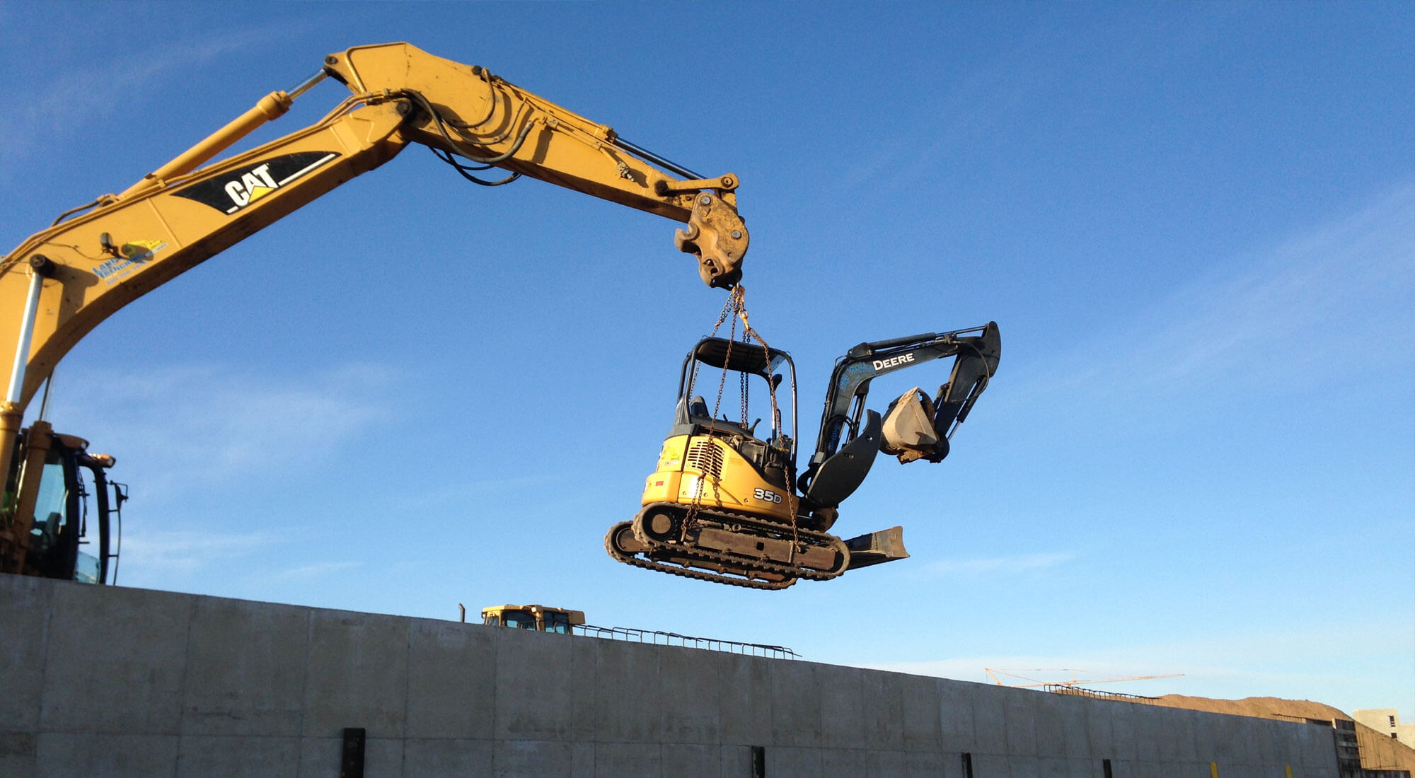 Large excavator lifting a mini excavator over a concrete wall